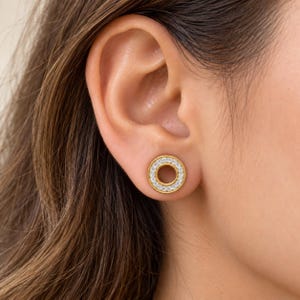 May include: A circular stud earring featuring a gold-toned metal frame with a central hollow opening. The outer ring is encrusted with a series of small, clear, sparkling stones arranged in a continuous circle, creating a bright, reflective surface.