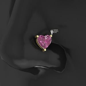 May include: A heart-shaped pink gemstone nose stud with a gold setting. The stud is set against a dark background. The jewelry is from Shirish Sparkle Gems.