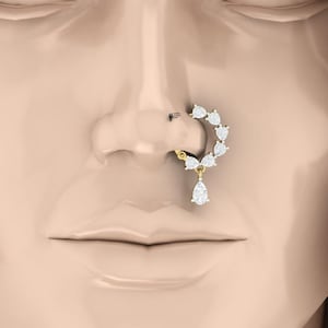 Op de afbeelding: Een gouden neusring met een gebogen rij van zes ronde witte stenen en een enkele peervormige witte steen als hanger. Het sieraad is geplaatst aan de zijkant van een neus en toont een fijne metalen band en heldere, geslepen witte stenen.