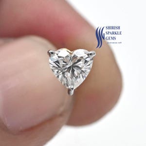 May include: A close-up of a heart-shaped diamond stud earring. The diamond is clear and faceted, with a brilliant sparkle. The earring is set in a silver or white gold setting. The text "SHIRISH SPARKLE GEMS" is visible.