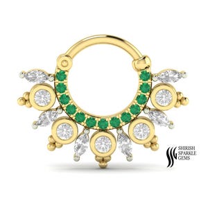 May include: A gold septum ring with a sunburst design. The ring features a row of small green gemstones and is accented with round and marquise-cut clear gemstones. The brand name "SHIRISH SPARKLE GEMS" is in the lower right corner.