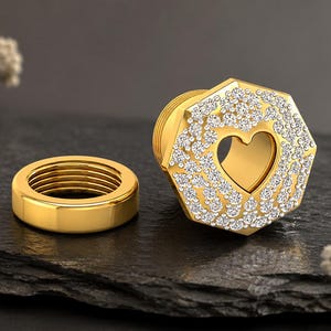 Può includere: Un dilatatore per orecchie color oro con una faccia ottagonale decorata con numerose piccole pietre chiare e scintillanti che circondano un ritaglio centrale a forma di cuore, accompagnato da una parte posteriore filettata.