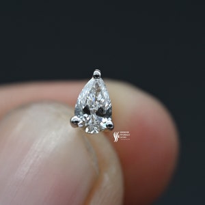 May include: A pear-shaped diamond gemstone set in a silver prong setting. The diamond is clear and sparkly.
