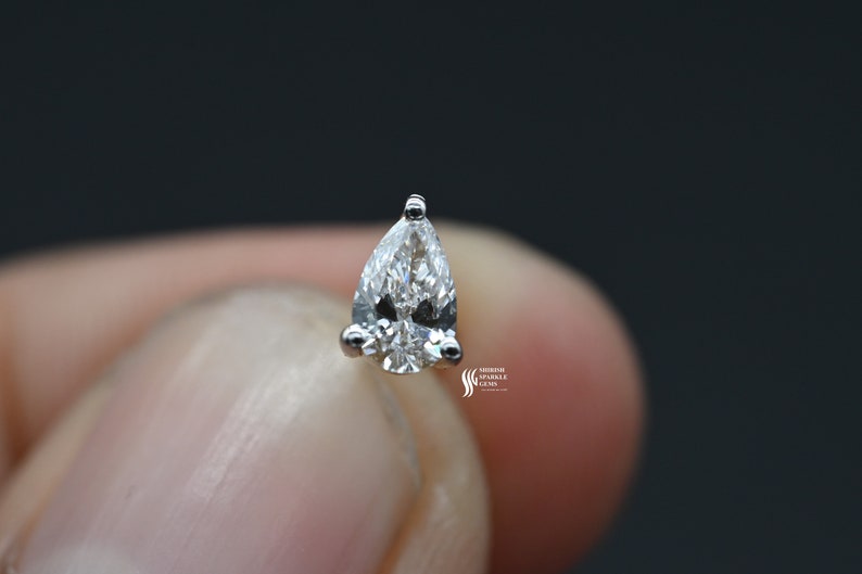 May include: A pear-shaped diamond gemstone set in a silver prong setting. The diamond is clear and sparkly.