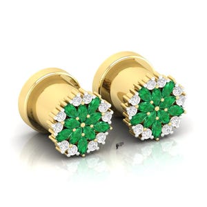 May include: A pair of gold ear plugs featuring a circular face adorned with a central cluster of marquise-cut green gemstones arranged in a floral pattern, surrounded by a border of small, round, clear white stones set in gold.