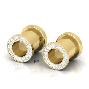 May include: A pair of gold-toned ear tunnels featuring a circular front face adorned with a ring of clear, baguette-cut stones. The cylindrical tunnels have a smooth, polished finish and a hollow center. The text "SWISH SPARKLE GEMS" is visible.