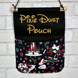 May include: A black fabric wall hanging pouch with a wooden dowel and black string. The top flap features the gold glitter text Pixie Dust Pouch. The lower pocket displays a holiday pattern with cartoon mice, a castle, and a white rabbit.