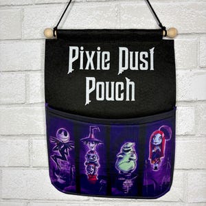 May include: A black felt wall hanging pouch with a wooden dowel and black cord for suspension. The text Pixie Dust Pouch is printed in white. The lower pocket features a purple graphic print of four characters from The Nightmare Before Christmas.