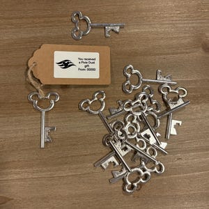 May include: A collection of silver-toned Mickey Mouse shaped keys with a bottle opener on the end. The keys are arranged in a pile on a wooden surface. A small brown tag with the text "You received a Pixie Dust gift From: 00000" is visible in the background.