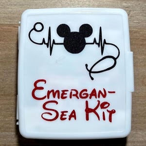 May include: White rectangular case with a black Mickey Mouse head and heartbeat line design. The text "Emergan-Sea Kit" is in red script. The case has a hinged lid and a rounded shape.