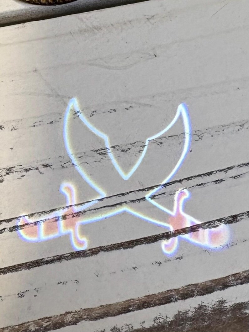 May include: An image of a crossed swords design, with a stylized, glowing effect. The swords are pale blue and pink, with a white outline, set against a white and brown wooden background.