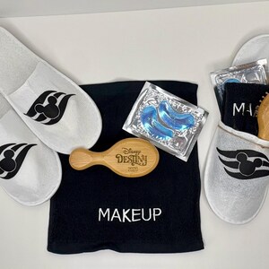 May include: A set featuring two white slippers with black Mickey Mouse icons, a black towel with the word MAKEUP embroidered in white, a wooden hairbrush engraved with Disney DESTINY 2025, and a packet of two blue under-eye gel patches.