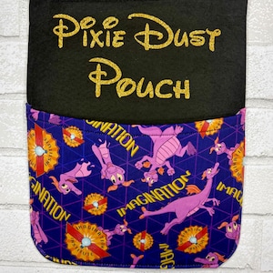 May include: A black felt wall hanging pouch with the gold glitter text Pixie Dust Pouch. The lower pocket features a purple fabric with a repeating pattern of pink dragons, yellow text reading Imagination, and orange circular Imagination Institute logos.