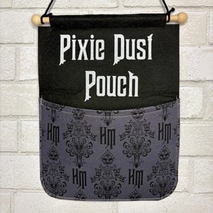 May include: A black fabric wall hanging with a pocket featuring a grey damask pattern and the letters HM. The top section is black with the white text Pixie Dust Pouch. It is suspended by a black cord from a wooden dowel with rounded ends.