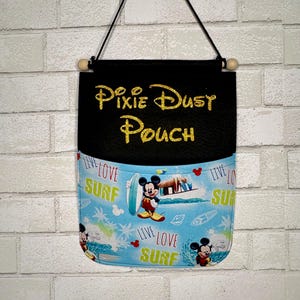 May include: A black fabric wall pouch featuring the gold glitter text Pixie Dust Pouch. The lower section is light blue with a repeating pattern of Mickey Mouse, surfboards, palm trees, and the text Live Love Surf, suspended by a black cord.