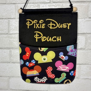 May include: A black fabric wall hanging pouch featuring the gold glitter text Pixie Dust Pouch. The lower pocket section displays a black background with a colorful pattern of various cartoon-style mouse ear hats and headwear designs.
