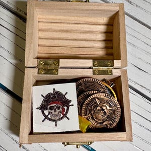 May include: A small, open wooden treasure chest with brass hinges and clasp. Inside are several gold-colored pirate coins and a temporary tattoo featuring a skull wearing a pirate hat.
