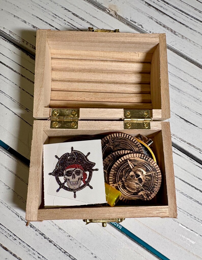 May include: A small, open wooden treasure chest with brass hinges and clasp. Inside are several gold-colored pirate coins and a temporary tattoo featuring a skull wearing a pirate hat.