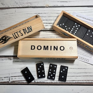 May include: Wooden domino set with black dominoes and white dots. The set includes three wooden boxes, two with the text "LET'S PLAY" and one with "DOMINO". A sheet of paper with rules is also included.