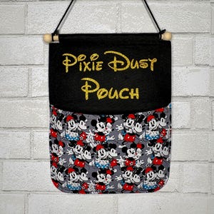 May include: A hanging fabric pouch featuring a black top section with the gold glitter text Pixie Dust Pouch and a grey bottom section patterned with a repeating print of Mickey and Minnie Mouse characters, suspended by a black cord and wooden rod.