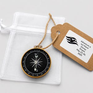 May include: A gold and black compass with a white star design, attached to a brown tag with the text "Adventure is out there! Disney Dream May 23-26, 2025". The compass is next to a white mesh bag.