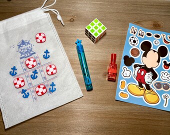 May include: A collection of toys and accessories on a wooden surface. Includes a white drawstring bag with a tic-tac-toe design, a Rubik's cube, a bubble wand, a small red container, and a sheet of Mickey Mouse stickers.