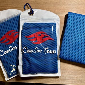 May include: Three blue cooling towels are displayed. Two are in clear plastic pouches with a white border and a blue carabiner clip. Each pouch has a red Mickey Mouse-shaped logo and the words "Cooling Towel" in white. The third towel is unfolded.
