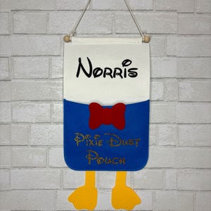 May include: A felt wall hanging featuring a design with the name "Norris" in black script above a blue pouch. The pouch has a red bow tie and the words "Pixie Dust Pouch" in gold script. Yellow feet hang from the bottom.
