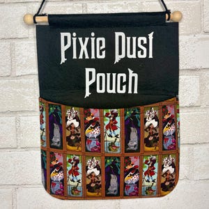 May include: A black fabric wall hanging pouch featuring the white text Pixie Dust Pouch at the top. The lower section displays a repeating grid pattern of colorful illustrations depicting various animated villain characters in rectangular frames.