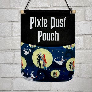May include: A black fabric wall hanging pouch featuring the white text Pixie Dust Pouch. The lower pocket section displays a dark blue pattern with yellow moons, stars, and cartoon characters including a tall skeleton and a ragdoll figure.