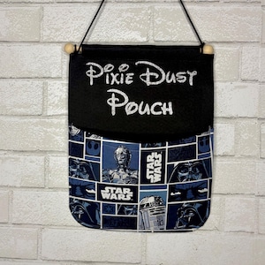 May include: A hanging fabric pouch featuring a black top section with the silver glitter text Pixie Dust Pouch and a bottom section with a blue and white Star Wars comic book style print including C-3PO, R2-D2, and Darth Vader imagery.