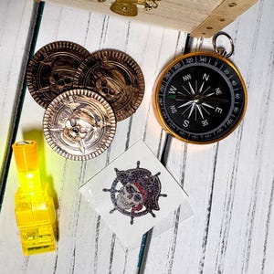 May include: A collection of pirate-themed items including three gold coins with skull designs, a compass, a yellow light, and a temporary tattoo with a skull and crossbones. The items are arranged on a white wooden surface.