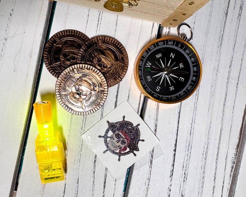 May include: A collection of pirate-themed items including three gold coins with skull designs, a compass, a yellow light, and a temporary tattoo with a skull and crossbones. The items are arranged on a white wooden surface.