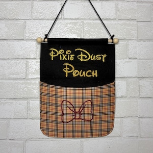 May include: A black fabric wall hanging pouch featuring the gold glitter text Pixie Dust Pouch at the top. The lower section consists of a tan, black, and red plaid fabric pocket decorated with a red glitter outline of a bow shape.