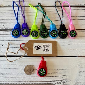 May include: A collection of colorful compass keychains with attached lanyards. The compasses are in various colors, including blue, purple, green, black, and pink. A tag with text that reads "Adventure is out there! Disney Dream May 23-26, 2025" is also present.
