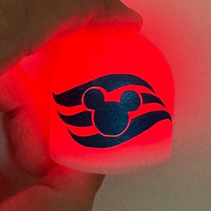 May include: A glowing red, cylindrical night light featuring a dark blue Mickey Mouse silhouette and stylized waves. The light is held in a hand, and the design is illuminated against a soft, white background.