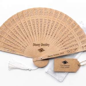 May include: A light brown wooden fan with intricate cut-out designs. The fan is open, displaying the words "Disney Destiny" and "Disney Destiny 2025". A small tag with the words "Fish Extender" and a logo is attached.