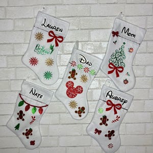 May include: Five white Christmas stockings with personalized names, each decorated with festive designs like gingerbread men, snowflakes, and bows. The names include "Lauren", "Dad", "Mom", "Nate", and "Aubrey".