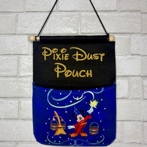 May include: A hanging wall pouch featuring a black felt top section with the gold glitter text Pixie Dust Pouch. The bottom section is blue fabric with a graphic of a cartoon mouse in a wizard robe, a broom, and two wooden buckets.