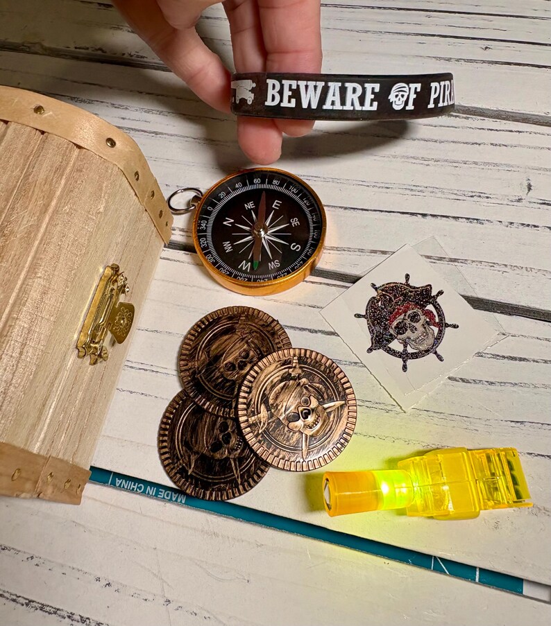 May include: A pirate-themed collection featuring a treasure chest, compass, coins, and a rubber bracelet that reads "BEWARE OF PIRATES". Includes a temporary tattoo and a small, yellow, glowing light.