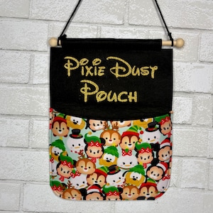 May include: A hanging wall organizer featuring a black felt top section with the gold glitter text "Pixie Dust Pouch" and a bottom pocket section patterned with a colorful print of various cartoon characters wearing holiday-themed accessories.