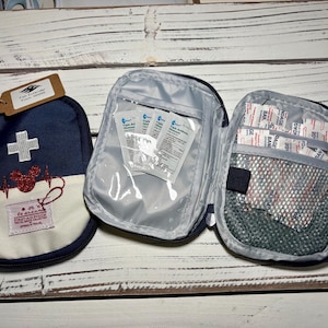 May include: Three navy blue and white first aid pouches. One pouch has a white cross and a red heart-shaped design. The other two pouches are open, revealing medical supplies. The text "be dreamin' FIRST AID POUCH" is visible.
