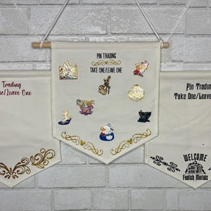 May include: Three off-white fabric banners for pin trading, each suspended by a wooden dowel and white cord. The banners display the text "Pin Trading Take One/Leave One" and decorative gold and black accents, with various pins attached.