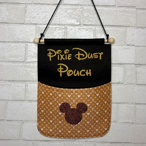May include: A hanging wall pouch featuring a black top section with the gold glitter text Pixie Dust Pouch and a tan bottom pocket with a white repeating pattern of small mouse silhouettes and a large dark brown glitter mouse head in the center.