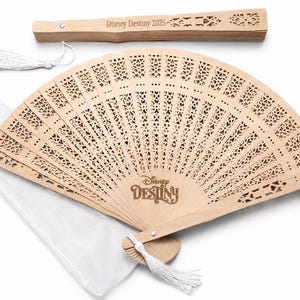 May include: A light-colored wooden fan with intricate cut-out designs, the words "Disney Destiny" are printed on the fan. A matching handle and a white tassel are attached. A white fabric pouch is also included.