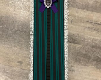 May include: A green and black striped fabric wall organiser with a purple bow and a white lace trim. The organiser has a label that says "Welcome to the Haunted Mansion".