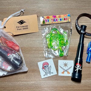 May include: A collection of pirate-themed party favors. Includes a drawstring bag with a pirate-themed item, a green plastic toy, a black telescope, a blue toy, a brown bracelet with text, and pirate-themed stickers. A tag reads "Fish Extender".