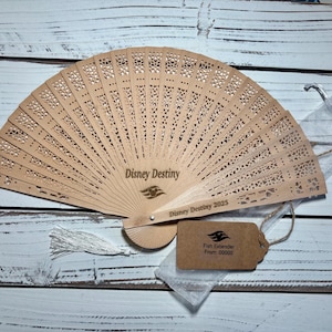 May include: A light brown wooden fan with intricate cut-out designs. The fan is open, displaying the text "Disney Destiny" and "Disney Destiny 2025". A small tag reads "Fish Extender From: 00000".