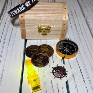 May include: A pirate-themed collection featuring a wooden treasure chest, gold coins, a compass, a glow stick, a temporary tattoo, and a bracelet with the text "BEWARE OF PIRATES".