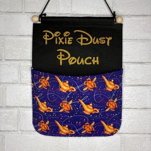 May include: A black fabric wall hanging pouch with the gold glitter text Pixie Dust Pouch. The lower pocket features a dark purple pattern with orange genie lamps and golden scarab beetles connected by thin, swirling white lines.